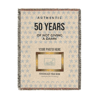 A rectangular woven throw blanket with fringe edges, featuring a beige background with a light blue floral pattern, black text reading 'AUTHENTIC 50 YEARS OF NOT GIVING A DAMN', a photo placeholder box, and smaller text 'PERSONALIZE YOUR BOOK'.