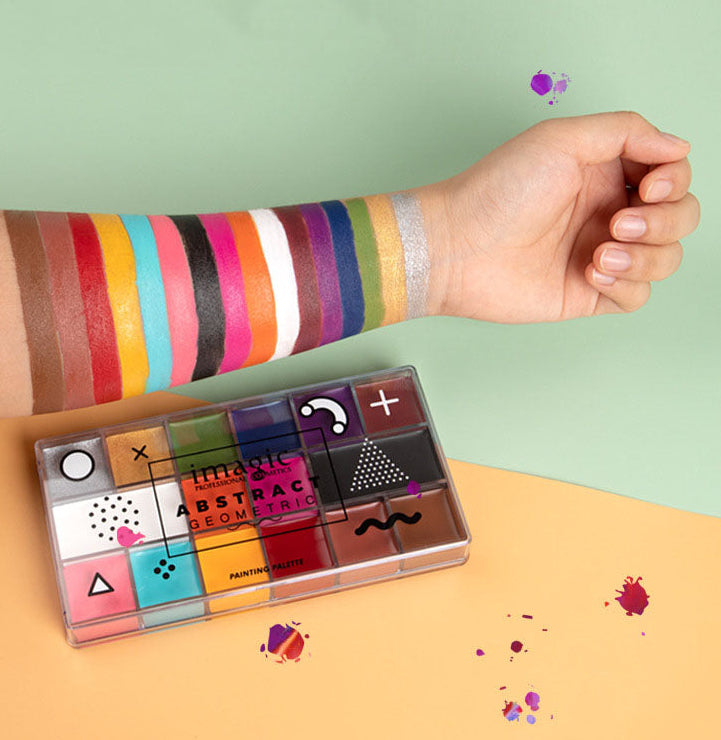 A clear rectangular palette labeled 'HISTORIC ABSTRACT' with multiple colorful paint wells, placed on a peach surface. A person's forearm and hand are visible across the frame, wearing or showing painted color stripes on the arm.