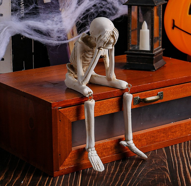 A Halloween skeleton figurine sitting on a wooden chest with its legs dangling down, styled with spooky props like fake cobwebs and a candle lantern.