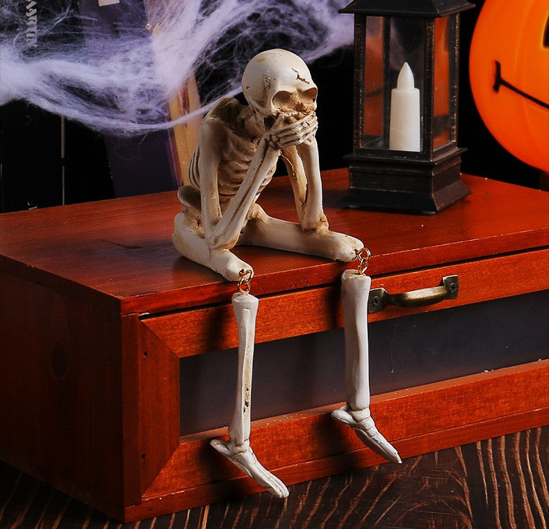 A posed skeleton decoration sitting on a reddish-brown wooden chest, with its legs dangling over the front. The scene includes Halloween-themed props, including fake cobwebs, a black lantern with a white candle, and part of an orange jack-o'-lantern in the background.