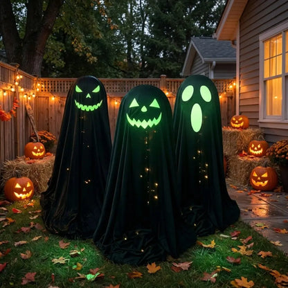 Three Halloween ghost decorations draped in black fabric stand on grass in a backyard yard, each with glowing green faces and small string lights, surrounded by pumpkins and fall leaves.