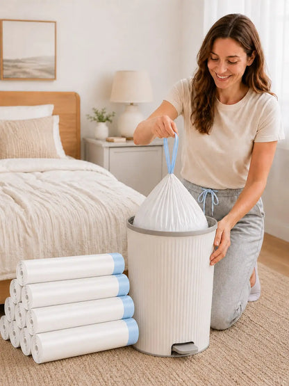 A woman kneels on a carpeted floor in a bright bedroom, placing a white plastic trash bag into a white ribbed cylindrical trash can with a gray foot pedal. Rolls of matching white trash bags with blue edges are stacked beside the can.