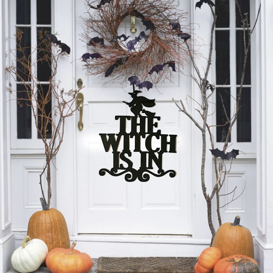 A black metal Halloween door sign reading 'THE WITCH IS IN' with a witch silhouette, displayed on a white front door surrounded by autumn decorations including pumpkins, a bat wreath, and bare branches.