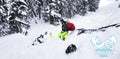 Two people in avalanche safety gear with a backpack, skiing down a snowy mountain slope.